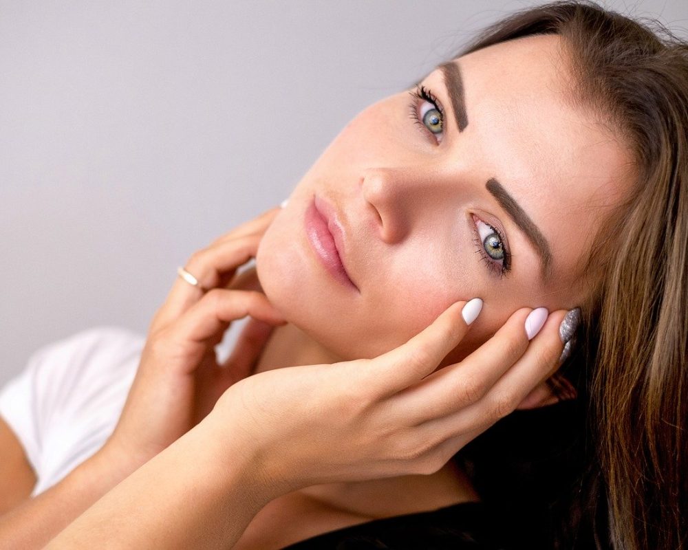 Woman gently touching her face, highlighting glowing skin and well-groomed eyebrows.